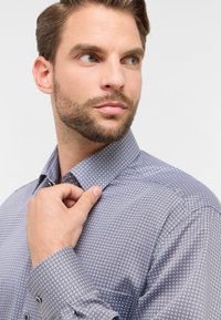 Camicia a fantasia con una base azzurro chiaro, caratterizzata da piccoli disegni geometrici. Ha un colletto e polsini abbottonati, realizzata in un tessuto morbido.