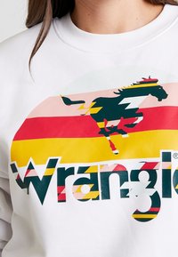 Primo piano di una persona che indossa una felpa bianca con un cavallo al galoppo multicolore e il logo "Wrangler" sul petto.