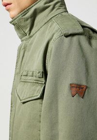 Olivgrön jacka med hög krage, axelkappar och två bröstfickor; har en brun läderpatch med Wrangler-logotyp.