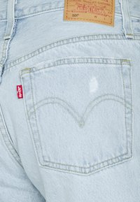 Los vaqueros de mezclilla de color azul claro presentan un bolsillo trasero con costuras visibles, una etiqueta roja de Levi's y una pequeña área desgastada encima del bolsillo.