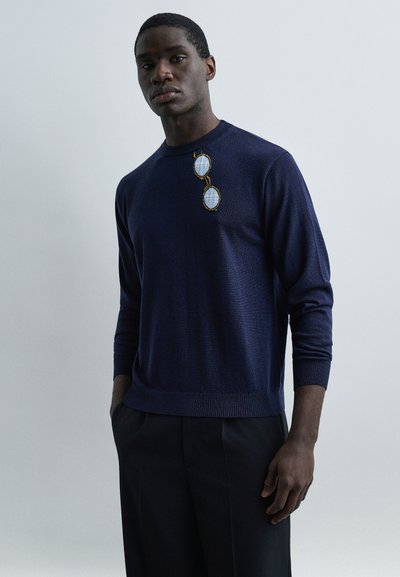Homme portant un pull bleu marine avec un motif de lunettes de soleil bleues brodé sur la poitrine, associé à un pantalon foncé, sur un fond uni.