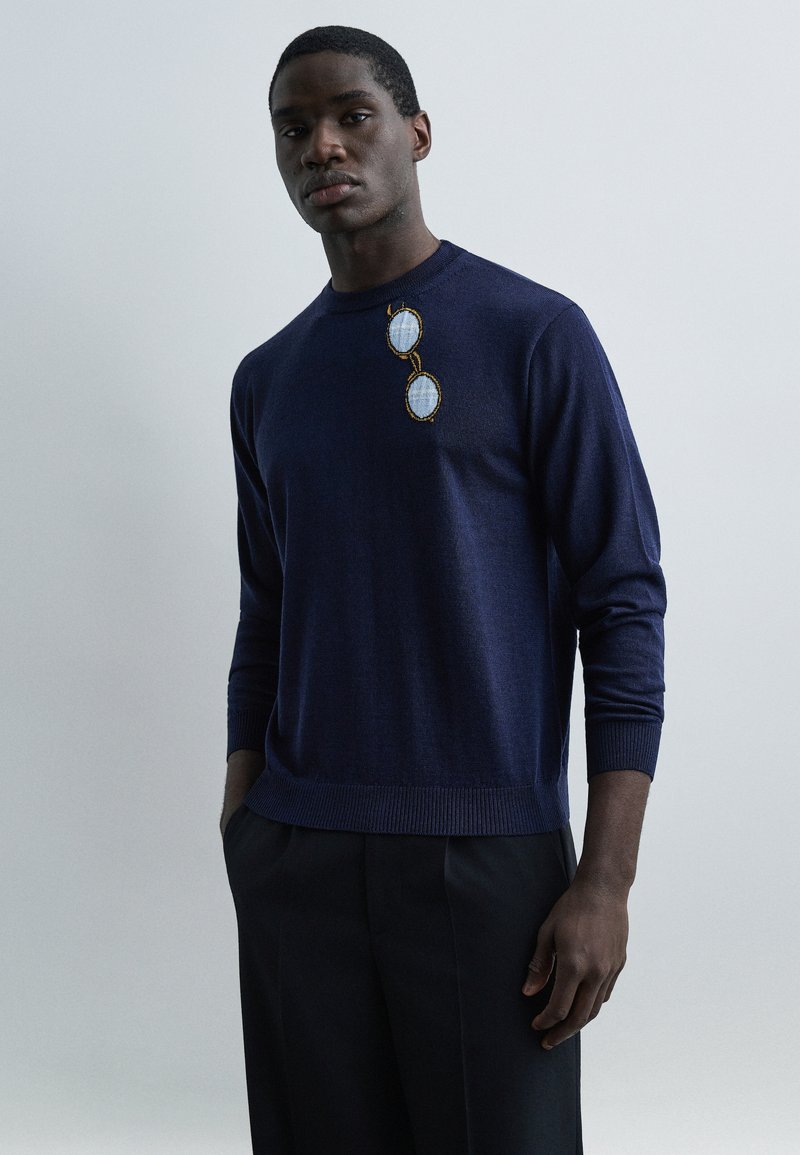 Άνδρας που φοράει μπλε σκούρο πουλ όβερ με κεντημένο σχέδιο μπλε γυαλιών ηλίου στο στήθος, συνδυασμένο με σκούρο παντελόνι, μπροστά σε απλό φόντο.