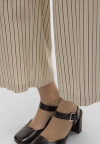 Piedi che indossano scarpe nere con tacco a blocco e cinturini alla caviglia, abbinate a pantaloni beige con sottili righe verticali nere.
