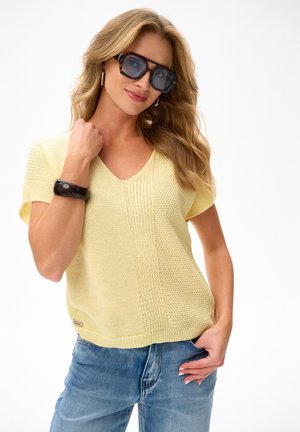 Vrouw met grote donkere zonnebril, geel kortmouwig gebreid shirt, blauwe jeans, zwarte armband aan linkerpols, poserend met hand in de zak.