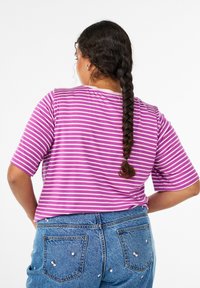 Pikkade punutud juustega naine kannab lillat ja valget triibulist särki ning siniseid teksaseid, millel on väikesed roosa kirsiga tikandid, seistes käed puusadel.