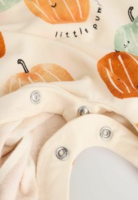 Tissu crème avec des motifs de citrouilles orange et verte, doté de boutons-pression argentés en bas. Texture douce, conçu pour les nourrissons.