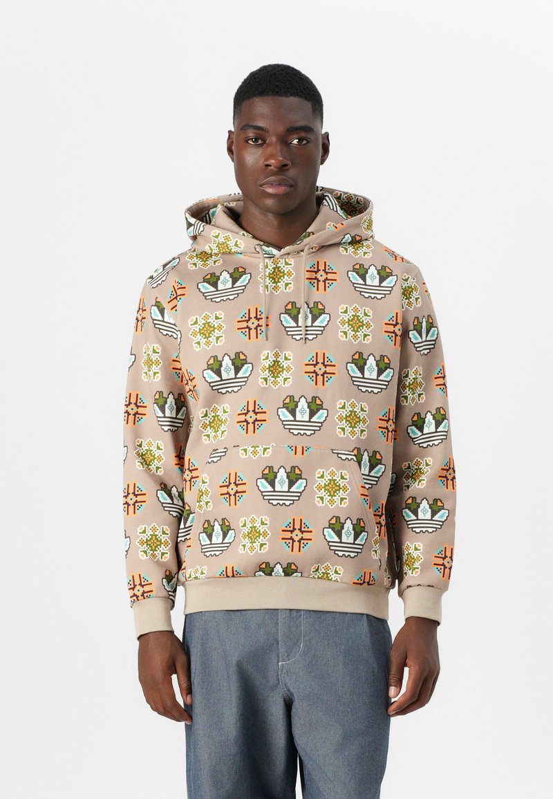 Giovane uomo che indossa un felpa beige con motivi geometrici e floreali colorati, abbinata a pantaloni blu larghi, in piedi contro uno sfondo bianco.