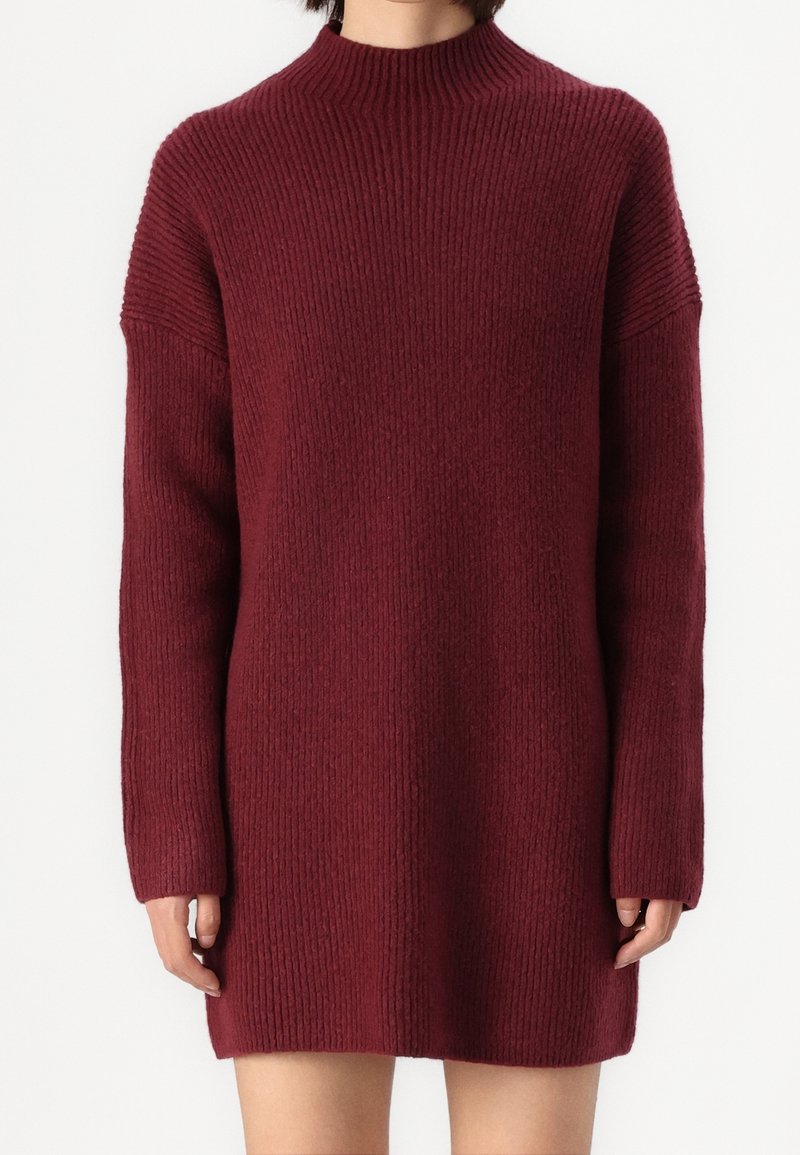 Robe-pull bordeaux à manches longues en maille côtelée avec col montant, portée par une personne montrée des épaules jusqu'à mi-cuisse sur un fond uni.