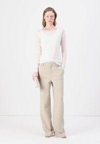 Lett, gjennomsiktig hvit langermet topp kombinert med beige vide bukser; modellen holder en liten beige håndveske. Enkelt og rent design.