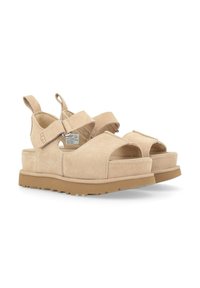 Sandales en suede beige avec orteils ouverts, sangles ajustables et une épaisse semelle en caoutchouc marron. Comprend le logo UGG sur le côté et un détail métallique.