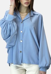 Lyseblå bluse med knapper foran, lange pufærmer og en glat satin tekstur. Har en klassisk krave og guldfarvede knapper.