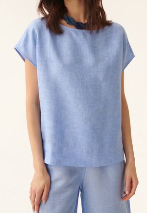 Femme portant un haut bleu clair à manches courtes et un pantalon assorti, accessoirisée d'un collier torsadé bleu foncé.