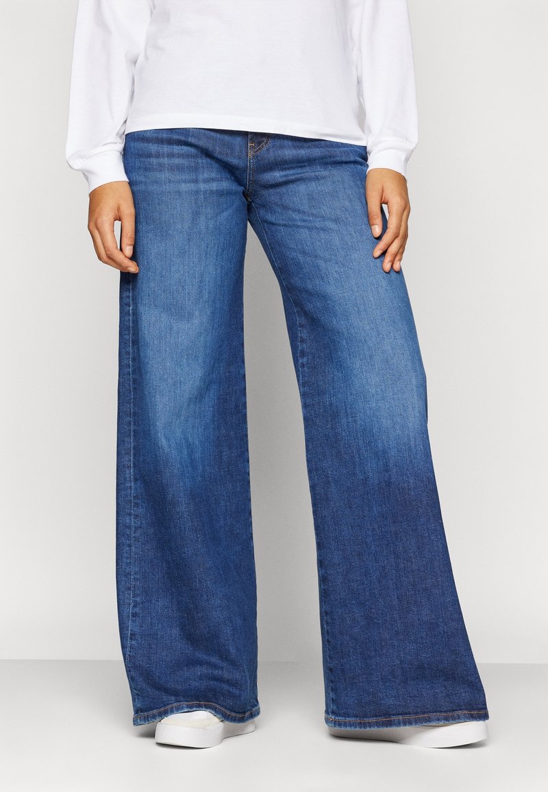Personne portant un jean bleu à jambes larges, une chemise blanche à manches longues et des baskets blanches, debout, les mains relâchées sur les côtés.
