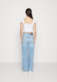 Kvinna med långt mörkt hår, iklädd en vit kort topp med blå blommönster, ljusblå hög midja vid byxor och vita sneakers, stående.