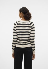 Gestreepte trui in zwart en crème, met een ronde halslijn en rib gebreide manchetten. Gezeigt op een model die zwarte broeken draagt.