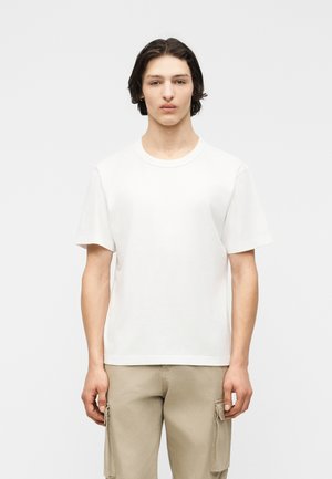 Mladý muž so stredne dlhými tmavými vlasmi, oblečený v jednofarebnom bielom tričku s krátkym rukávom a béžových nohaviciach typu cargo, stojaci na neutrálnom pozadí.