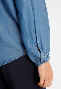 Vaaleansininen chambray pitkähihaine paita, jossa on nappikiinnityksellä varustetut hihansuut ja rypytetty yksityiskohta ranteissa. Sujuva tekstuuri, rento mitta.