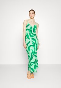 Vestido verde em slip com um padrão de folhas onduladas, tecido leve, alças finas e uma bainha suave. Combinado com sandálias laranjas com tiras.