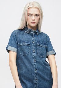 Jeansskjorte med korte rullede ermer, to brystlommer, knapping foran og en lys til middels blå farge med en lett tekstur.