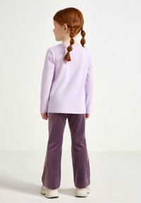 Chemise longue à manches avec col rond de couleur violet clair, associée à un pantalon évasé en velours violet. Cheveux tressés de l'enfant visibles de derrière.