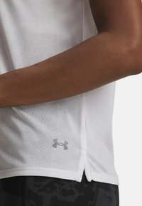 Vit mesh sportströja med en sidöppning, med en grå Under Armour-logotyp vid fållen. Slät textur och lätt design.