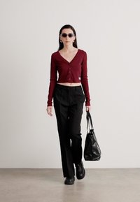 Cardigan scurt cu maneci lungi de culoare burgund, cu butoane, pantaloni negri croiți, geantă neagră și ochelari de soare întunecați; texturi netede și siluetă ajustată.