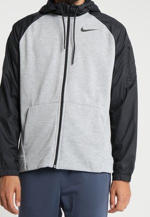 Muž nosící šedou mikinu na zip s černými rukávy a kapucí, s malým černým logem Nike na levé straně hrudi.