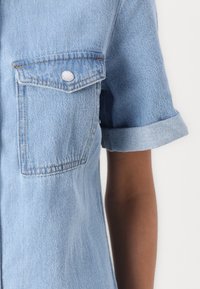 Lys blå denimskjorte med korte rullede ermer, to brystlommer og sølvfargede trykknapper. Har en klassisk, avslappet passform og teksturert stoff.