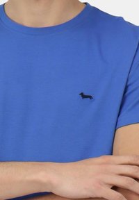 T-shirt in cotone blu con maniche corte, caratterizzato da un ricamo di un piccolo bassotto nero sulla parte sinistra del petto. Texture liscia.