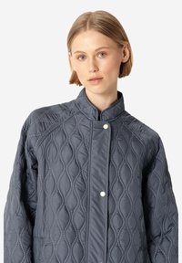 Chaqueta acolchada de color gris oscuro con cuello de pie, que presenta un patrón de diamantes, cierre a presión y detalles de costura en contraste.