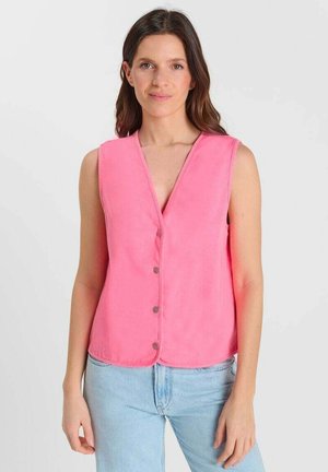 Donna con capelli castani lunghi che indossa un gilet rosa senza maniche con bottoni e jeans azzurri, in piedi contro uno sfondo bianco uniforme.