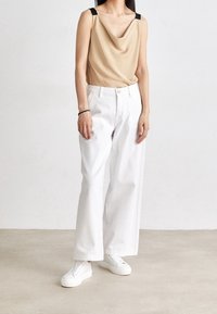 Beige topp med kappa och svarta axelremmar, ihop med löst sittande vita byxor och vita sneakers på en neutral bakgrund.