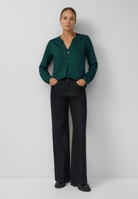 Ciemnozielona bluzka z wciętym kołnierzykiem i trzema guzikami, zestawiona z czarnymi spodniami o szerokich nogawkach i czarnymi butami. Gładka tekstura materiału.