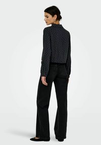 Femme portant un blouse noire à manches longues avec de petits imprimés roses et un jean large taille haute noir, se tenant de dos.