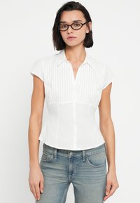 Νεαρή γυναίκα με κοντά σκουρόχρωμα μαλλιά και γυαλιά, που φοράει λευκό κοντομάνικο πουκάμισο με κουμπιά και μπλε τζιν, στέκεται μπροστά σε λευκό φόντο.