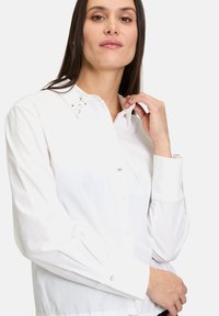 Camicia bianca con bottoni, adornata con cristalli decorativi sul colletto e sui polsini. Il materiale è liscio e con una leggera lucentezza, caratterizzata da una vestibilità classica.