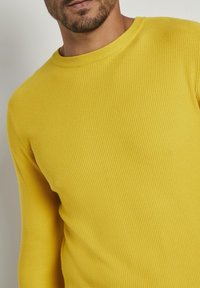 Żółty sweter prążkowany z okrągłym dekoltem i teksturowanym materiałem, ukazujący zbliżenie na szczegóły szycia i dopasowanie.
