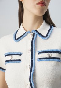 Bílá fleecová košile s krátkým rukávem, tmavě modré a světle modré pruhy, dvě přední kapsy a kovové knoflíky s květinovým vzorem.