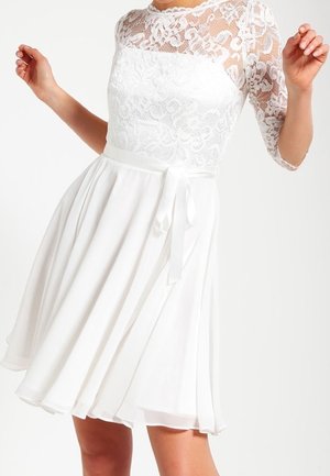 Vestido blanco hasta la rodilla con parte superior de encaje floral transparente, mangas tres cuartos y cinturón de satén atado, llevado por una persona de pie.