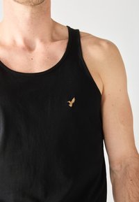 Camiseta sin mangas negra de algodón, con un cuello redondo y un logo bordado de un águila dorada en el lado izquierdo.