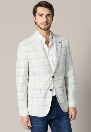 Mężczyzna w jasnoszarej marynarce z niebieskim wzorem okienka, białej koszuli i ciemnoniebieskich dżinsach, stojący na neutralnym tle.