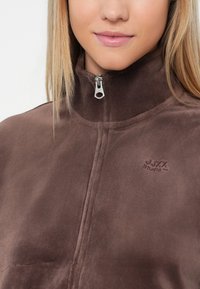 Smeđi flis pulover s visokim ovratnikom i četvrtim patentnim zatvaračem. Ima izvezeni logo na prsima, glatku teksturu i prilagođeni kroj.