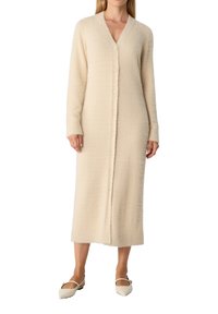 Cardigan lungo di colore crema realizzato in morbido tessuto testurizzato. Presenta un collo a V, chiusura con bottoni e maniche lunghe.