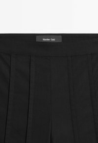 Zwarte broek van een gestructureerde stof, met voorplooien en een label dat aan de band is gestikt. Eenvoudig ontwerp zonder zichtbare hardware.