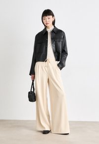 Svart lær cropped jakke med frontlommer, kremfarget blondertop, beige vide bukser og liten svart håndveske. Jevne teksturer generelt.