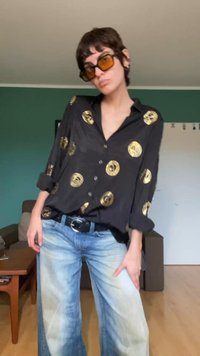 Black button-up shirt with golden circular patterns, long sleeves rolled at elbows. Paired with light blue wide-leg jeans and a black belt.