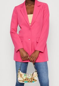 Blazer rose à deux boutons avec des revers crantés, associé à un sac à main à motifs avec des accents dorés et une bandoulière, porté avec un jean.