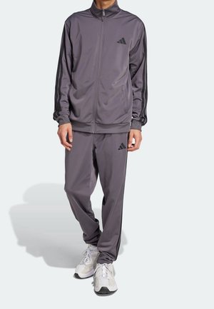 Mann trägt grauen Adidas-Trainingsanzug mit schwarzen Streifen und Logo, kombiniert mit weißen Sportschuhen, steht vor einem schlichten Hintergrund.