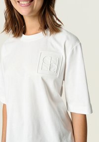 White cotton t-shirt with short sleeves, featuring a textured logo patch on the left chest. Smooth fabric with a classic fit.