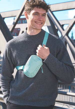 Borsa a tracolla azzurro chiaro con un motivo geometrico, con chiusura a zip e tracolla regolabile, indossata sopra un maglione in maglia grigio scuro.
